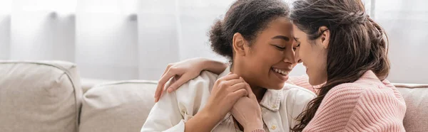 Positive lesbian woman hugging multiracial partner and sitting on couch, banner — Stock Photo