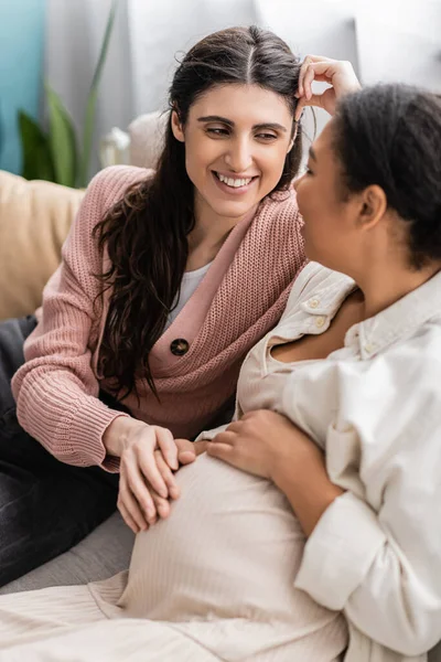 Happy lesbian woman touching tummy of pregnant multiracial partner and sitting on couch — Stock Photo