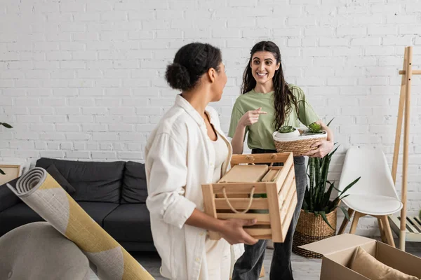 Happy lesbian woman pointing at wicker basket with plants near multiracial girlfriend during relocation to new house — Stock Photo