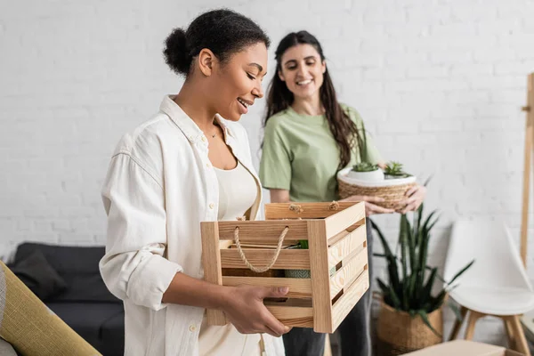 Mulher multirracial feliz segurando caixa de madeira perto do parceiro lésbico durante a mudança para a nova casa — Fotografia de Stock