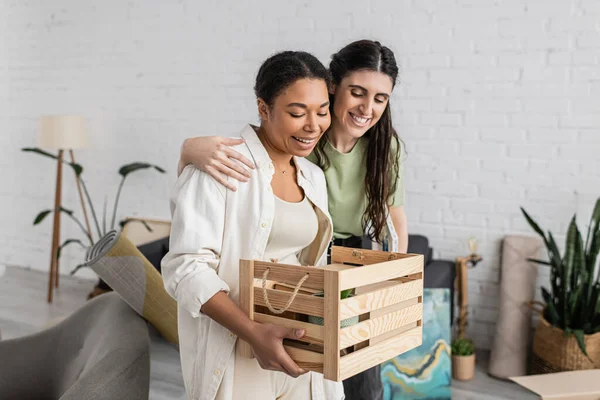 Overjoyed lesbian woman hugging multiracial girlfriend holding wooden box during relocation to new house — Stock Photo