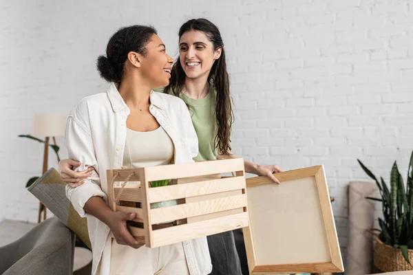 Mujer multirracial feliz sosteniendo caja de madera y mirando a la pareja lesbiana durante la reubicación a nueva casa - foto de stock