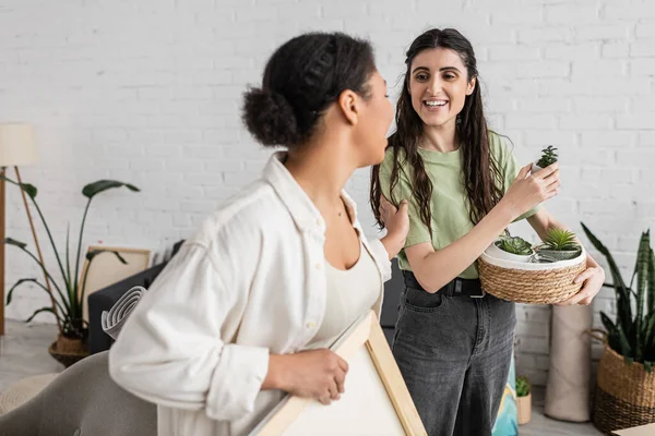Cheerful lesbian woman holding wicker basket with plants while looking at multiracial girlfriend during relocation to new house — Stock Photo