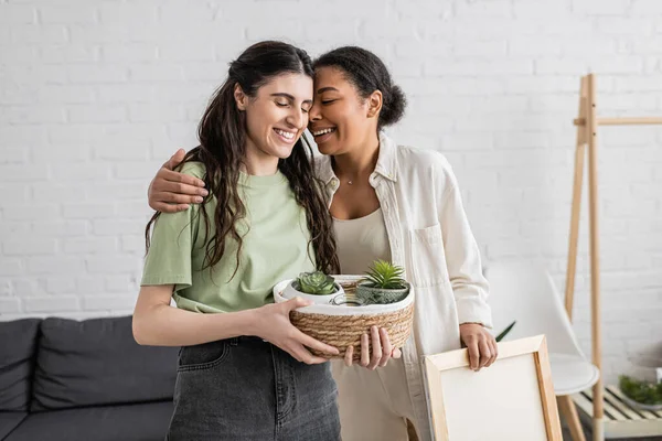 Happy multiracial woman hugging cheerful lesbian girlfriend holding wicker basket with plants in new house — Stock Photo