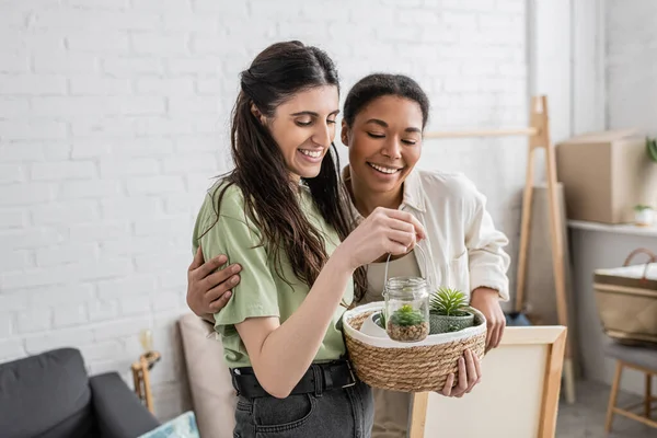 Fröhliche lesbische Frau hält Weidenkorb und Glasgefäß mit Pflanzen neben positiver multirassischer Freundin im neuen Haus — Stockfoto
