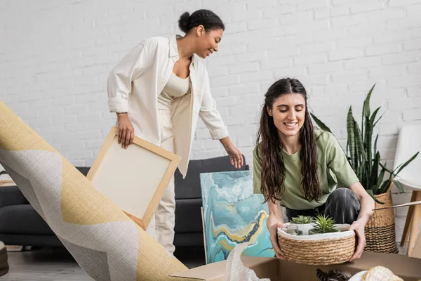 Cheerful lesbian woman holding wicker basket with green plants near multiracial girlfriend next to paintings in new house — Stock Photo