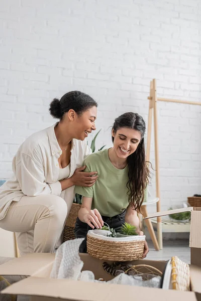 Happy lesbian woman looking at green plants near multiracial girlfriend in new house — Stock Photo