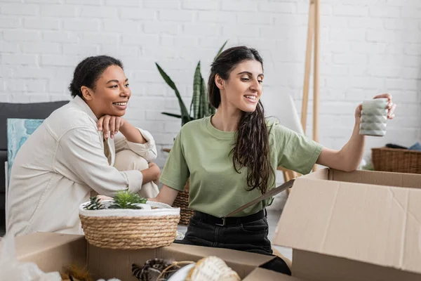 Heureuse lesbienne femme déballer bougie de la boîte près de petite amie multiraciale dans une nouvelle maison — Photo de stock