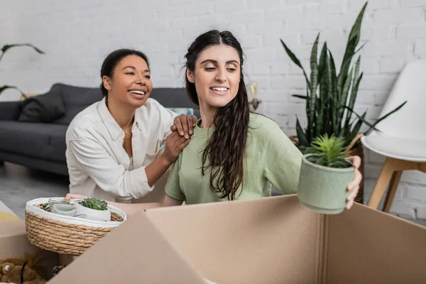 Happy lesbian woman unpacking plant in flowerpot near multiracial girlfriend in new house — Stock Photo
