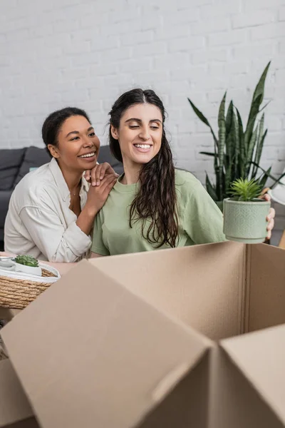Overjoyed mulher lésbica desempacotando planta em vaso perto de namorada multirracial na nova casa — Fotografia de Stock