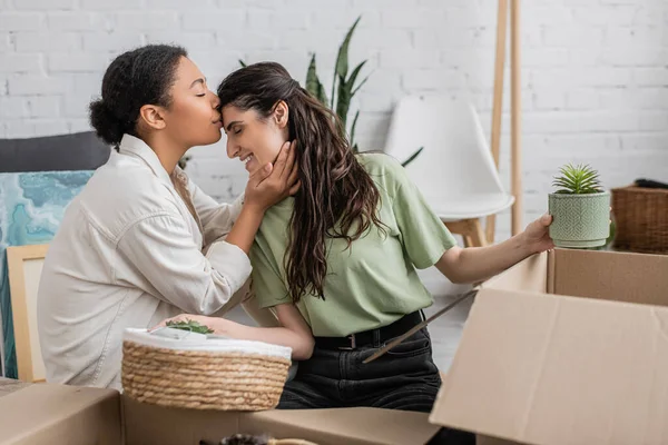 Joyful multiracial woman kissing forehead of happy girlfriend while moving to new house — Stock Photo