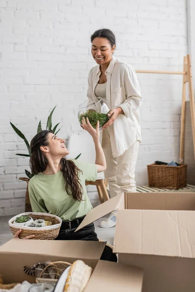 Joyeuse femme multiraciale tenant vase en verre avec plante verte près partenaire lesbienne lors de la relocalisation — Photo de stock