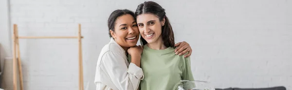 Mujer multirracial llena de alegría abrazando a su pareja lesbiana en una casa nueva, pancarta - foto de stock