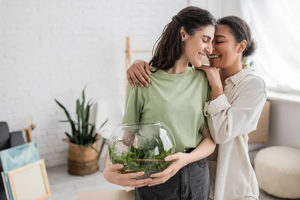 Позитивная многорасовая женщина обнимает лесбиянку-партнера с зеленым растением в стеклянной вазе — стоковое фото