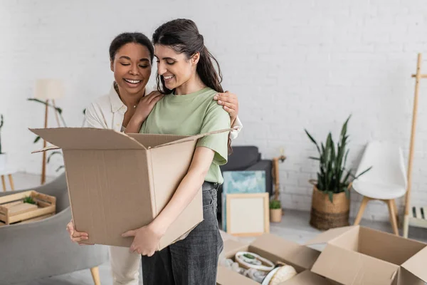 Positive multiracial woman hugging lesbian partner with carton box in hands — Stock Photo