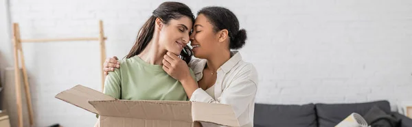 Mujer multirracial positiva abrazando a su pareja lesbiana con caja de cartón, pancarta - foto de stock