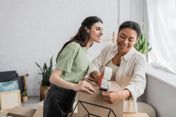 Cheerful multiracial woman holding tape dispenser near carton box and happy lesbian partner during relocation to new house — Stock Photo