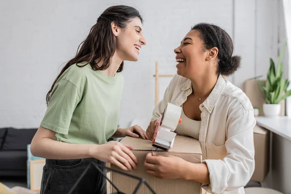 Joyful multiracial woman holding tape dispenser near carton box and happy lesbian partner during relocation to new house — Stock Photo