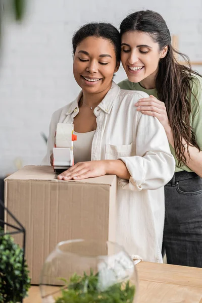 Femme souriante multiraciale tenant distributeur de ruban adhésif près de la boîte en carton et partenaire lesbienne heureux lors de la réinstallation dans une nouvelle maison — Photo de stock