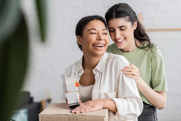 Alegre multirracial mujer celebración cinta dispensador cerca caja de cartón y riendo con lesbiana pareja - foto de stock