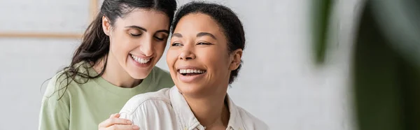 Alegre multirracial mujer riendo con lesbiana pareja en casa, bandera - foto de stock