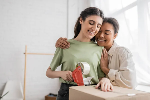 Cheerful multiracial woman hugging happy lesbian partner taping carton box during relocation to new house — Stock Photo