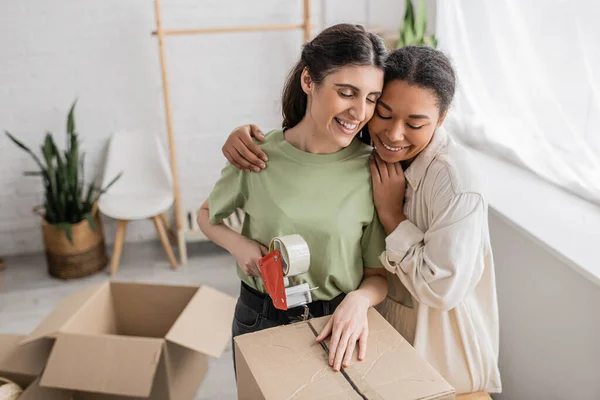 Sonriente mujer multirracial abrazando feliz pareja lesbiana grabando caja de cartón durante la reubicación a nueva casa - foto de stock