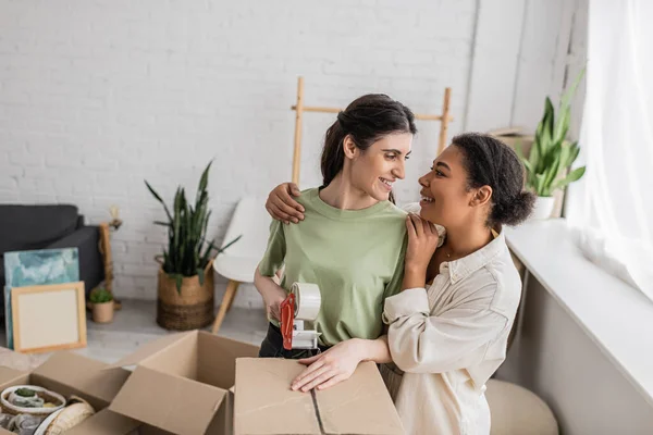Lesbiana multirracial mujer abrazando feliz novia cinta caja de cartón durante la reubicación a nuevo casa - foto de stock