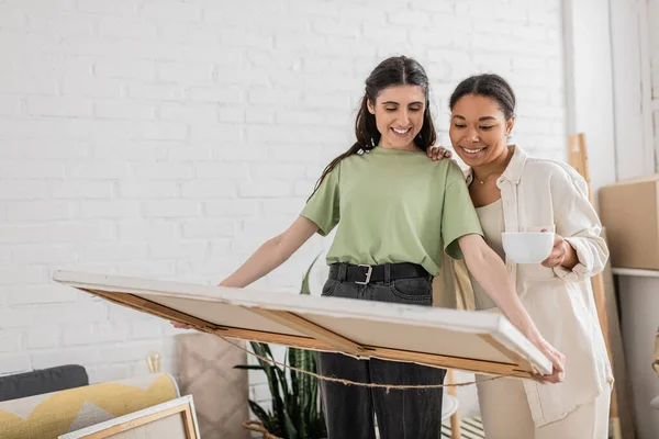 Cheerful interracial lgbt couple looking at painting while standing in new house — Stock Photo