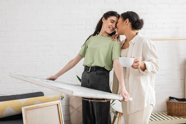 Happy lesbian woman with closed eyes holding painting near multiracial girlfriend with cup of coffee — Stock Photo