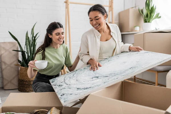 Cheerful interracial lgbt couple looking at painting while unpacking box in new house — Stock Photo
