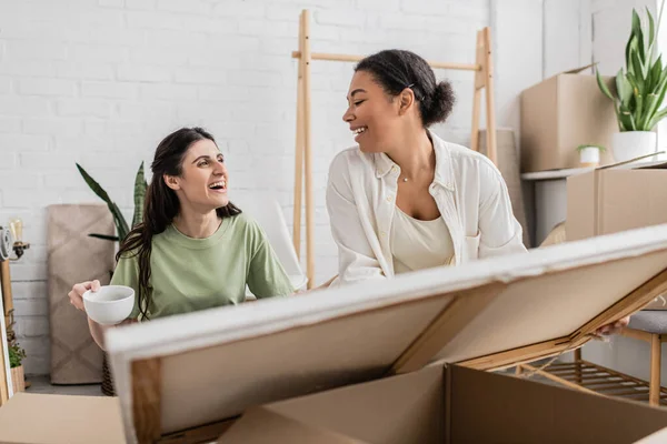 Happy interracial lesbian couple looking at each other while unpacking painting from box in new house — Stock Photo