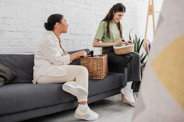 Joyful interracial lesbian couple holding wicker baskets while sitting on sofa in living room — Stock Photo