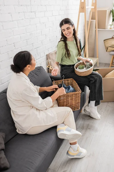 Vista aérea de feliz interracial lesbianas pareja sosteniendo cestas de mimbre mientras se sienta en sofá en sala de estar - foto de stock