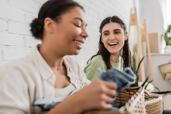 Mulher lésbica alegre olhando para namorada multirracial dobrar roupas em primeiro plano borrado — Fotografia de Stock