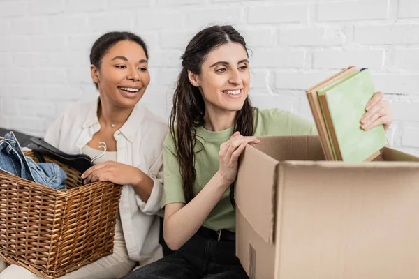 Joyful lesbian woman unpacking books from carton box while sitting on sofa next to multiracial girlfriend — Stock Photo