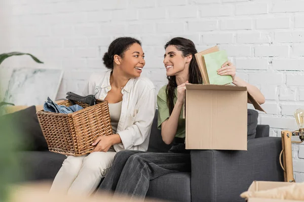 Happy lesbian woman unpacking books from carton box while sitting on sofa next to multiracial girlfriend — Stock Photo