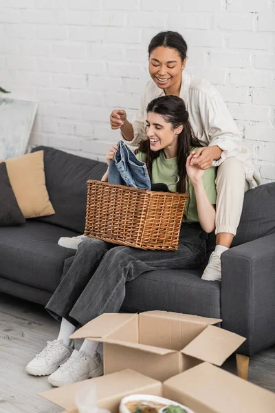 Alegre lésbicas mulher dobrar limpo roupa enquanto sentado no sofá ao lado de multirracial namorada — Fotografia de Stock