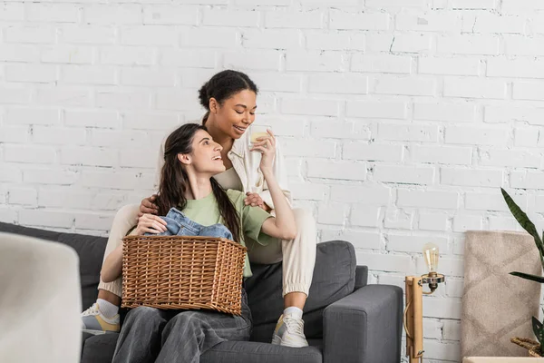 Joyful lesbian woman holding scented candle while sitting on sofa next to multiracial girlfriend — Stock Photo