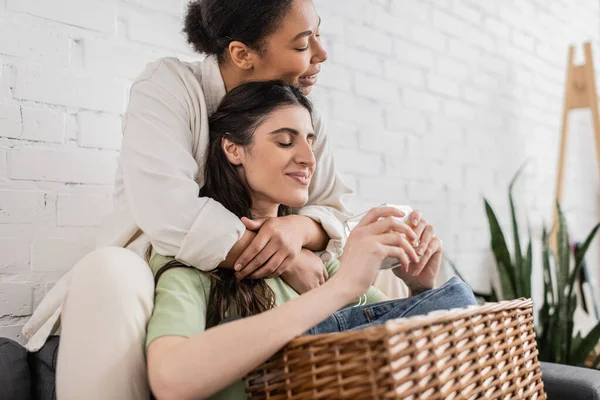 Joyful lesbian and multiracial woman hugging girlfriend holding scented candle — Stock Photo