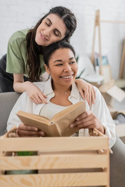 Cheerful lesbian woman hugging multiracial girlfriend with book in hands — Stock Photo