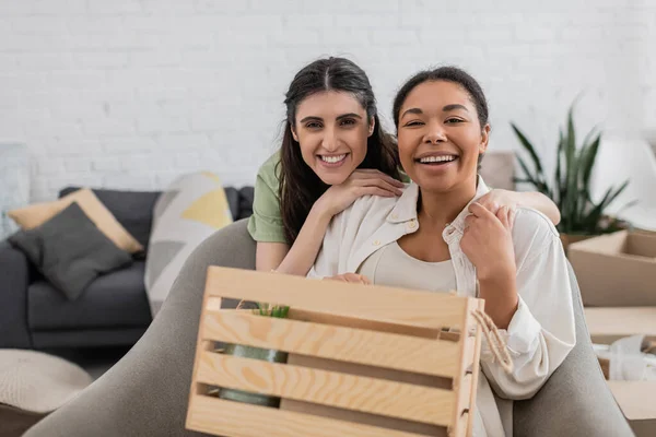 Alegre mulher lésbica abraçando feliz namorada multirracial perto de caixa de madeira na sala de estar — Fotografia de Stock
