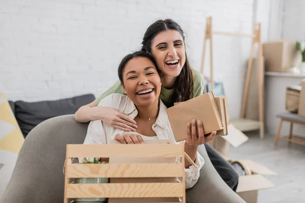 Fröhliche lesbische Frau umarmt glückliche multirassische Freundin mit Büchern im Wohnzimmer — Stockfoto