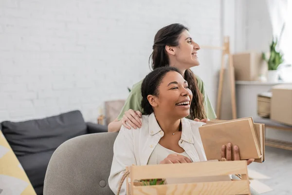 Positive lesbian woman hugging happy multiracial girlfriend with books in living room — Stock Photo