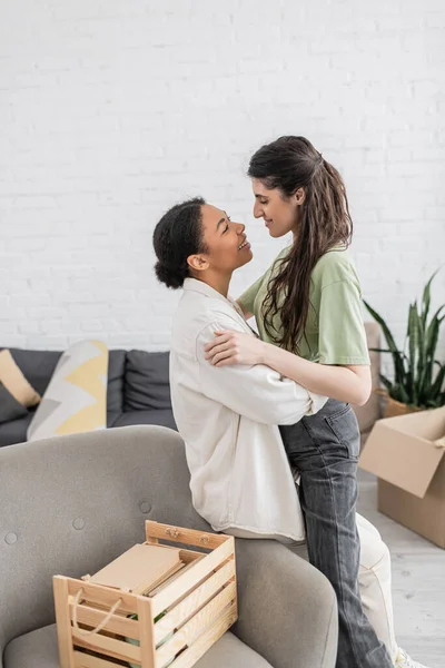 Mujer lesbiana positiva abrazando feliz novia multirracial cerca de caja de madera en la sala de estar - foto de stock