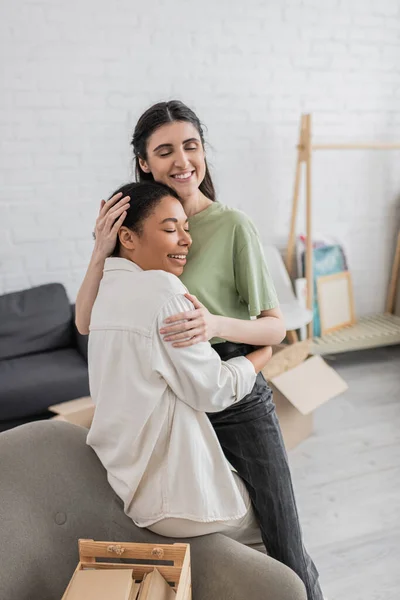 Zufriedene lesbische Frau umarmt glückliche multirassische Freundin mit geschlossenen Augen im Wohnzimmer — Stockfoto