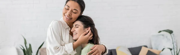 Compiaciuta donna lesbica che abbraccia gioiosa fidanzata multirazziale con gli occhi chiusi in soggiorno, banner — Foto stock