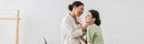 Donna lesbica sorridente che abbraccia gioiosa fidanzata multirazziale in soggiorno, banner — Foto stock
