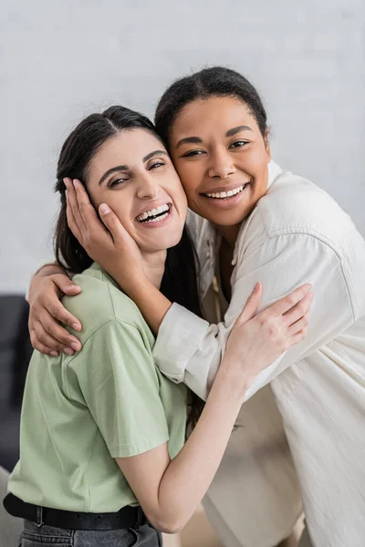 Fröhliche lesbische Frauen, die sich im Wohnzimmer umarmen und in die Kamera schauen — Stockfoto