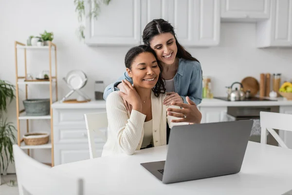 Счастливая межрасовая лесбийская пара смотрит на ноутбук, обнимаясь на кухне — стоковое фото